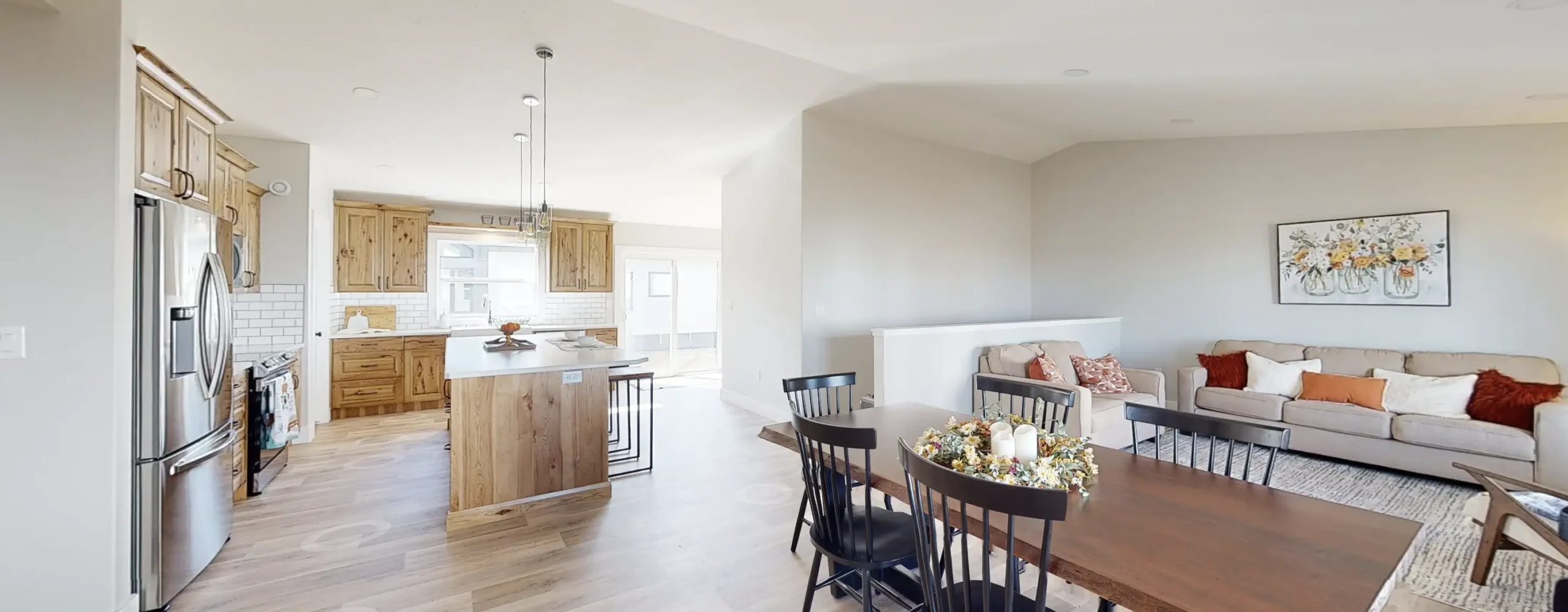 Open concept RTM home in Alberta or Saskatchewan with knotty wood kitchen cabinets and island, white subway tile, stainless appliances, black dining chairs, and a beige sofa.